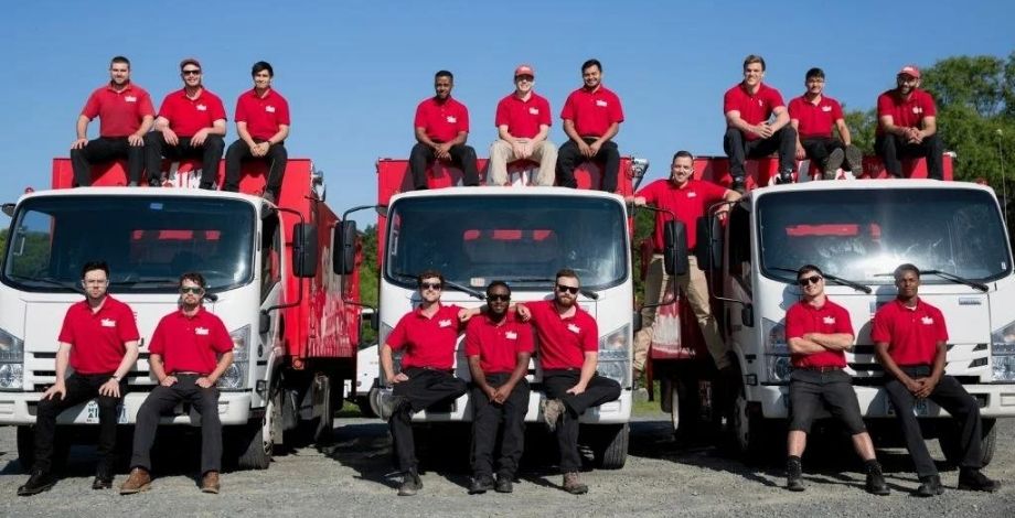 Group photo of 123JUNK crew members in red shirts posing on and in front of three junk removal trucks