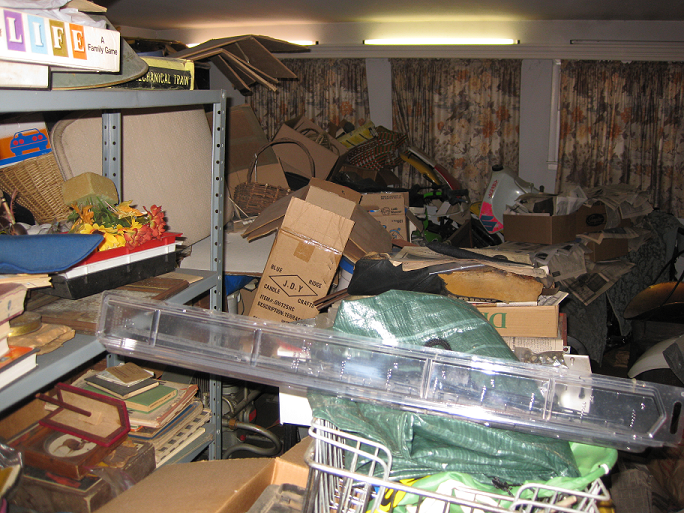 Cluttered living room filled with boxes, bags, baskets, and miscellaneous household items blocking furniture and surfaces