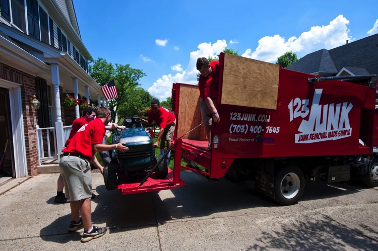 Team of workers in red shirts loading a riding lawn mower into a 123JUNK truck in a residential driveway