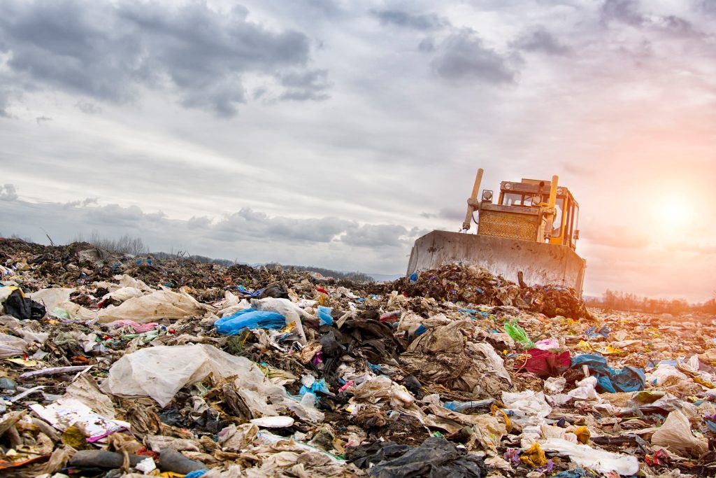 Landfill scene with a bulldozer pushing piles of mixed trash under a cloudy sky with sunlight breaking through