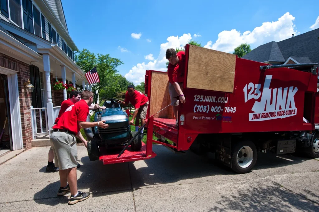 Team of workers in red shirts loading a riding lawn mower into a 123JUNK truck in a residential driveway