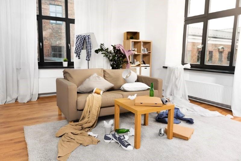 A messy living room with clothes, bottles, and other items scattered on the floor and furniture