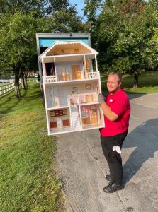 Junk removal worker in a red shirt carrying a large dollhouse outdoors toward a truck