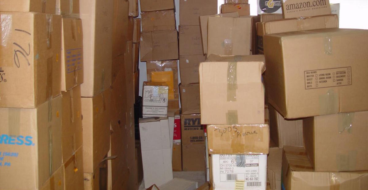 A room stacked high with numerous cardboard boxes labeled and sealed, creating narrow walking space