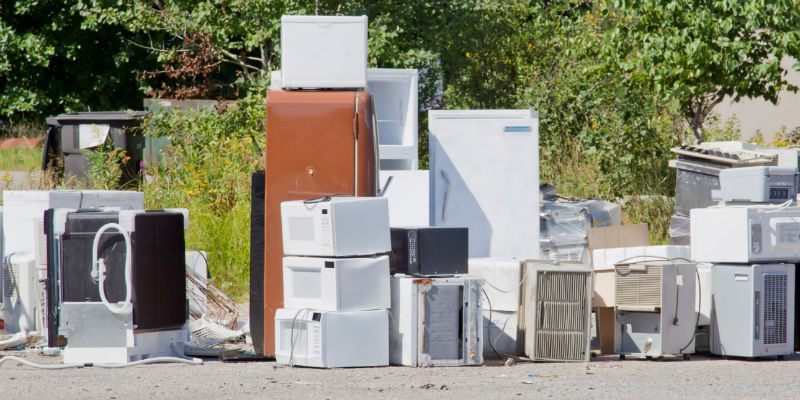 Outdoor scene showing a pile of discarded appliances including refrigerators, microwaves, and air conditioning units