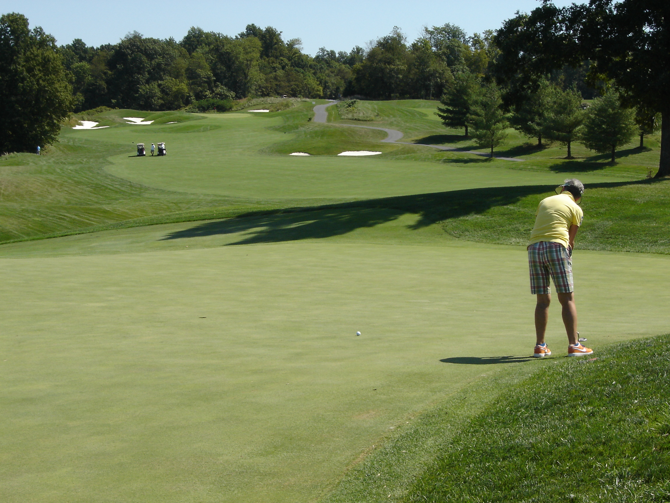 A golfer wearing a yellow shirt and plaid shorts putting on a green with sand traps and golf carts in the background