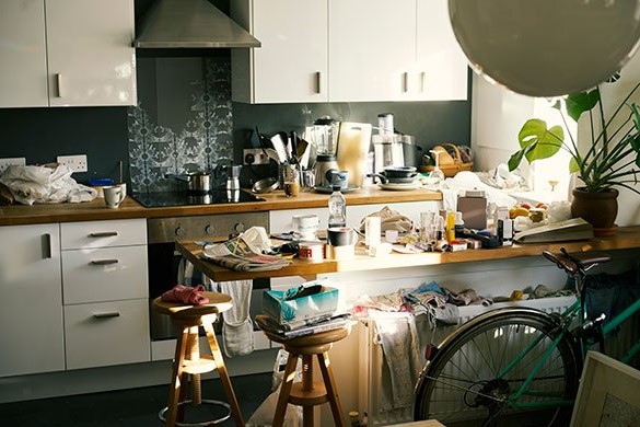 Cluttered kitchen with countertops and stools covered in various household items, and a bicycle partially visible in the foreground