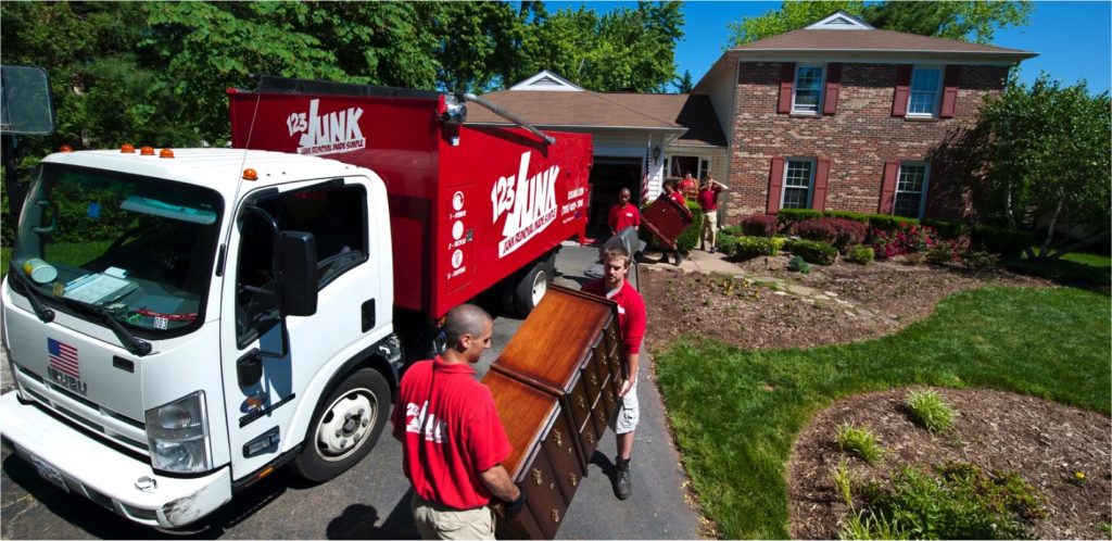 Junk removal crew in red shirts carrying furniture from a suburban house into a large red junk removal truck parked in the driveway