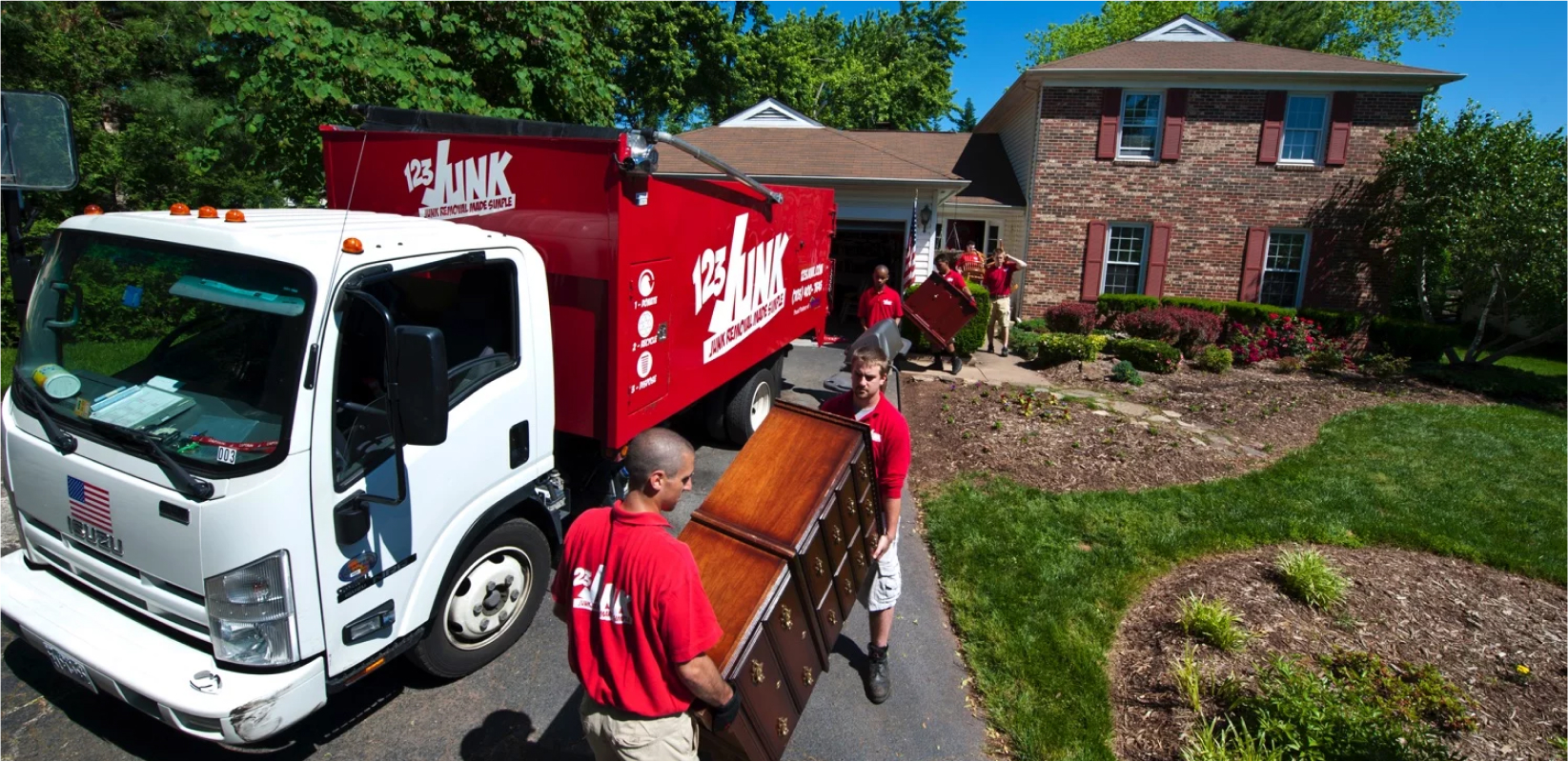 Junk removal crew in red shirts carrying furniture from a suburban house into a large red junk removal truck parked in the driveway