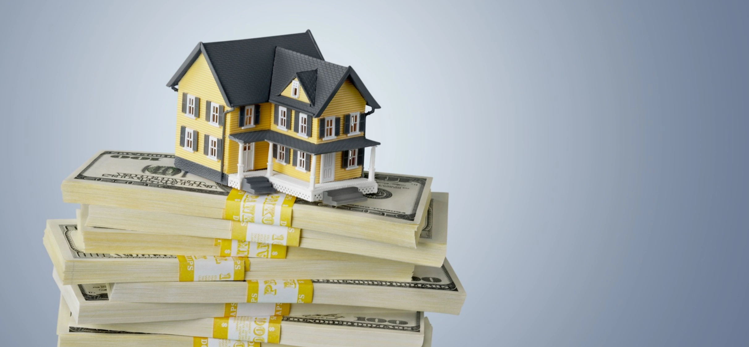 Model of a yellow house placed on top of several stacks of hundred-dollar bills against a blue background